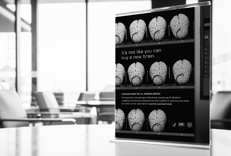 Black and white photo of Surgeon General print ad standing on a table in an office environment with desks in the background